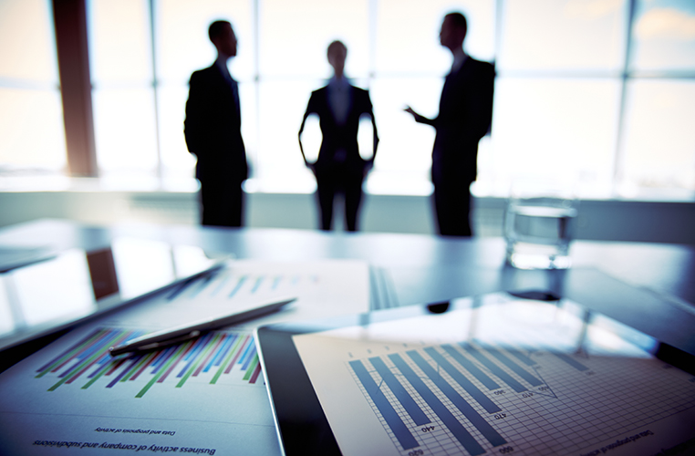 Focused reports on desk with three people standing blurred in background