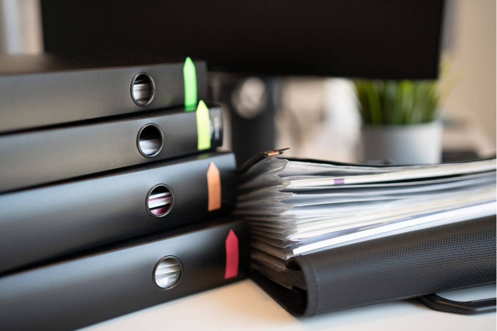 1. Stack of black binders with coloured tabs and a pile of documents on a desk, symbolising ISO certification and quality management documentation.